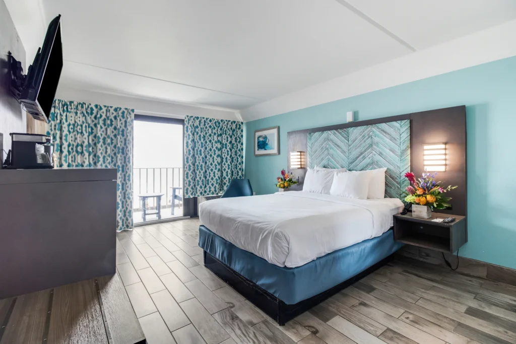 luxurious coastal themed bedroom with a single queen sized bed, washed grey wooden furniture, facing a large sea green accent wall, with patterned curtains in the background shading an oceanfront view