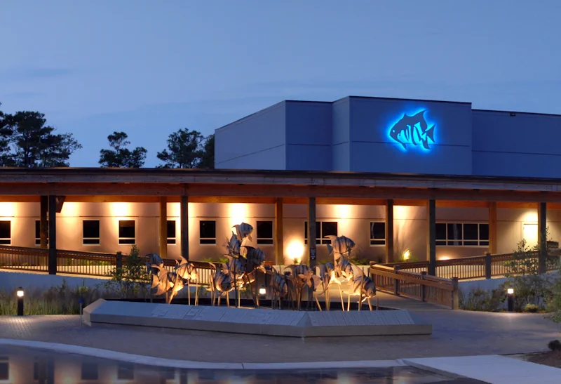 a nighttime photo of a large, wide building with a roofed pathway out front. on top of the building is a glowing blue fish logo. in front of the building is a metal sculpture of a school of fish.