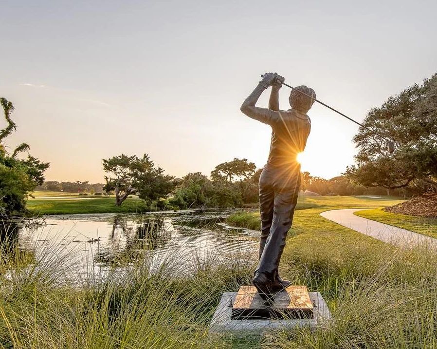 a stone statue of a golfer swinging a golf club, behind it are fields of green grass, a river of water, and a stone pathway leading into the distance.