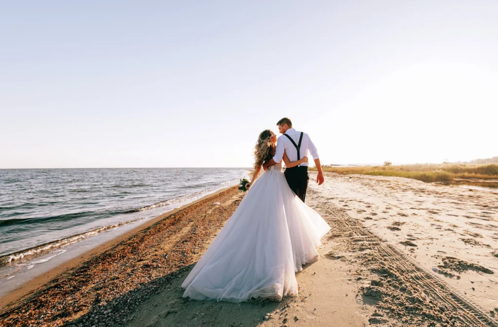 a handsome man in formal wear and a beautiful woman in a wedding dress holding a bouquet walk away from the camera down the crystal coast with the sun setting in the distance