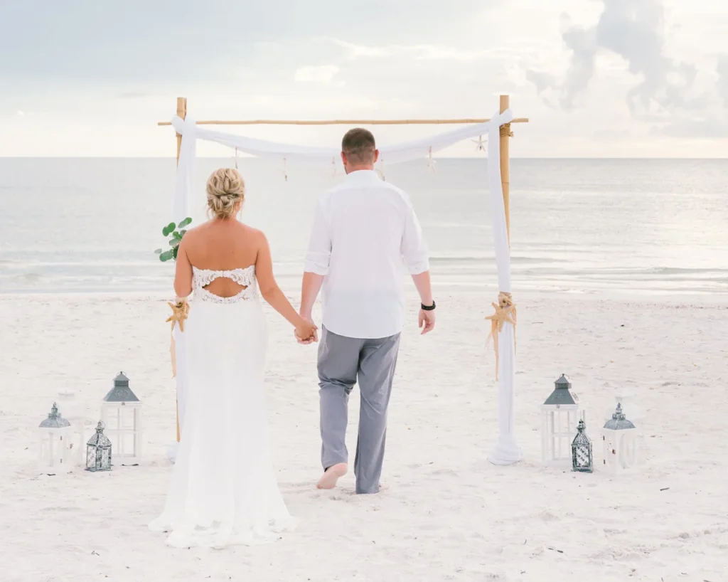 a brightly lit photo of a man in grey pants and a white shirt and a woman in a wedding dress holding hands and facing the ocean, with a white cloth archway between them and the water