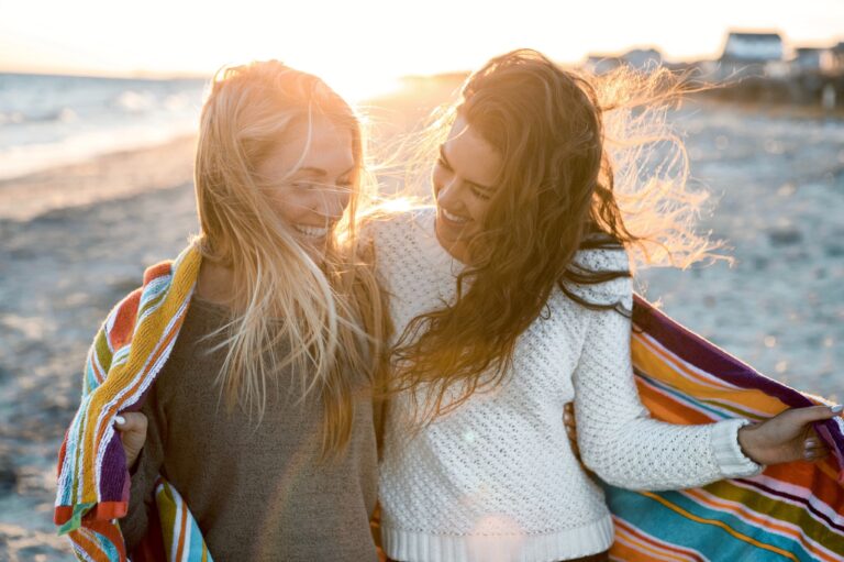 two girls laugh and smile at each other while sharing a towel and walking along the crystal coast shoreline at sunset
