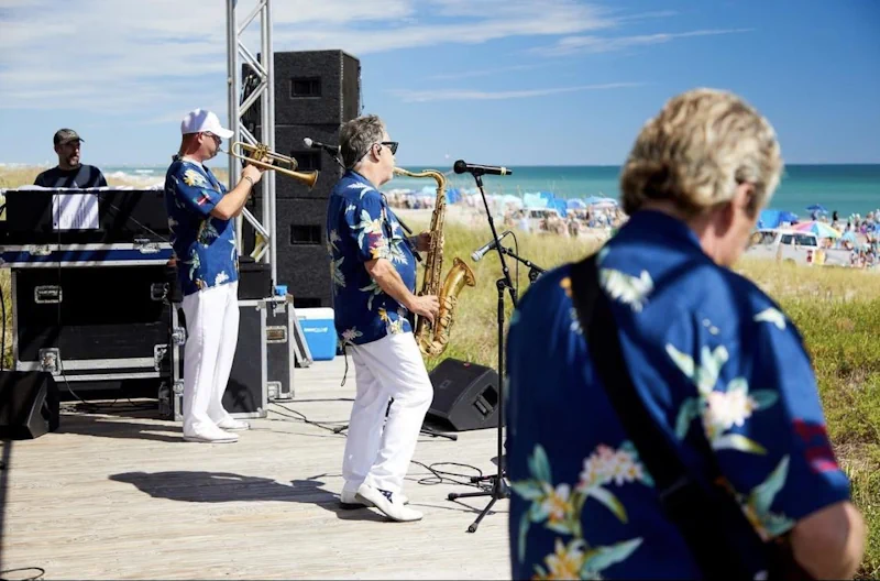 a band of men with various instruments plays to a beach full of umbrellas and people dancing along the crystal coast beach