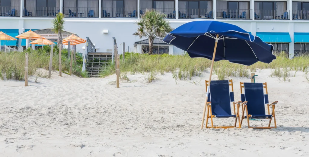 two navy blue chairs sit under an blue umbrella in the sand. in the background, a wooden boardwalk leads up to the crystal coast oceanfront hotel