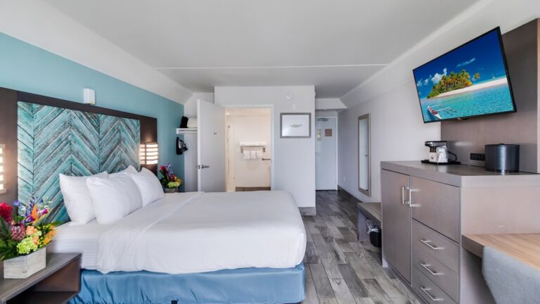 luxurious coastal themed bedroom with a queen sized bed, washed grey wooden furniture, and a sea green accent wall with a flatscreen TV across from the bed. an open door in the background reveals an all white bathroom