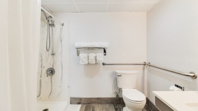 all white bathroom with white walls, a marble lined shower with silver polished shower pipes, ADA-compliant accessibility handrails, and a toilet.