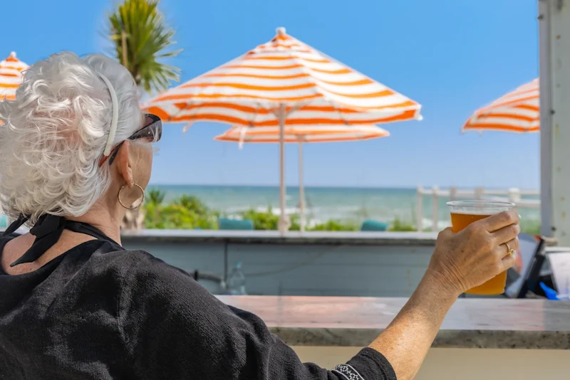 an older woman looks into the distance while holding up a glass of golden colored beer. in the background are orange umbrellas, a sandbar, and a path leading to the sea green waters of the beach