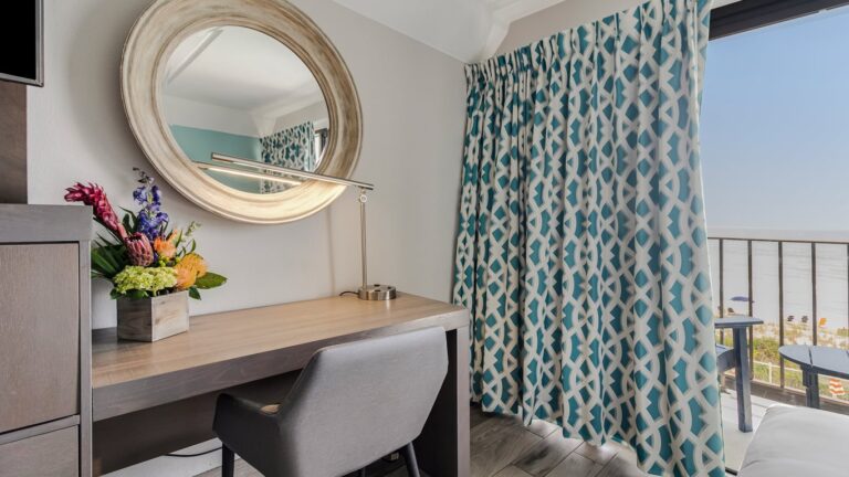 a washedout grey wooden desk with a large golden rimmed window sits near a sea green and white patterned curtain half open to reveal a balcony facing the crystal coast ocean