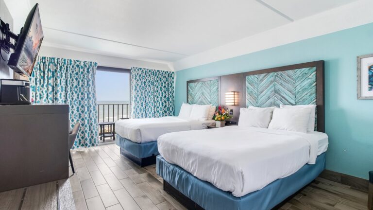 luxurious coastal themed bedroom with a two standard sized bed, washed grey wooden furniture, facing a large sea green accent wall, with patterned curtains in the background shading an oceanfront view