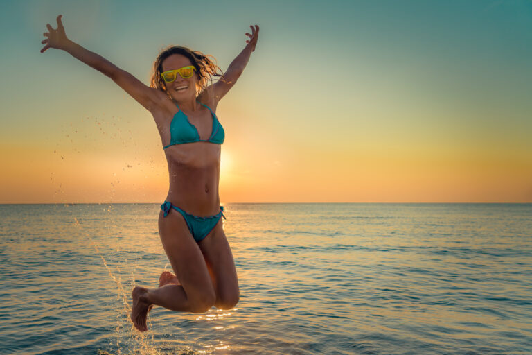 a woman jumping in the ocean shore water, facing the camera with a bright smile and wide open arms, sunset behind her