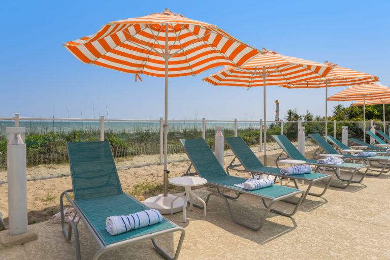 teal fabric grey metal chairs in a row sit under a series of orange umbrellas around a pool with the crystal coast in the background