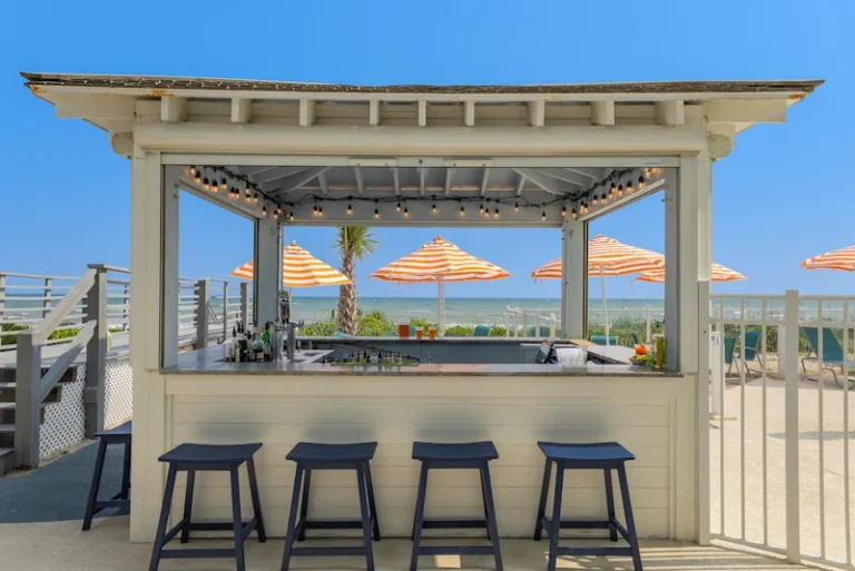 a white painted wooden shelter with open walls, hanging LED lights, navy blue barstools, and a variety of liquor options stands near a pool with the ocean in the background