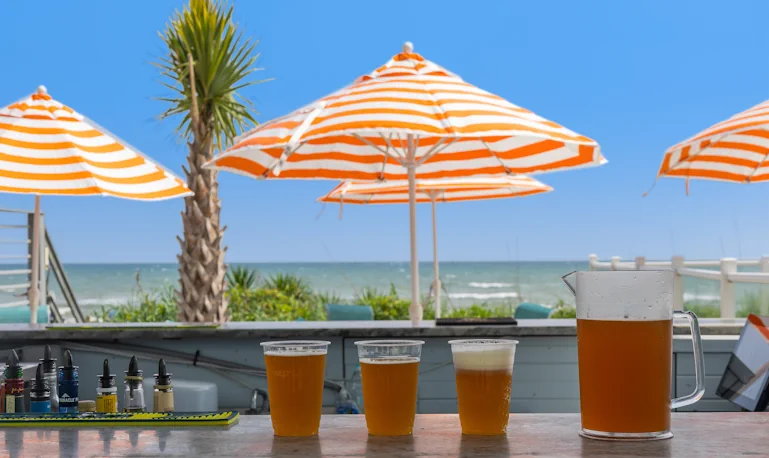 four frosty cold glasses of beer sit on a white marble counter in the foreground, with a beautiful sunny crystal coast with palm trees and orange umbrellas in the background