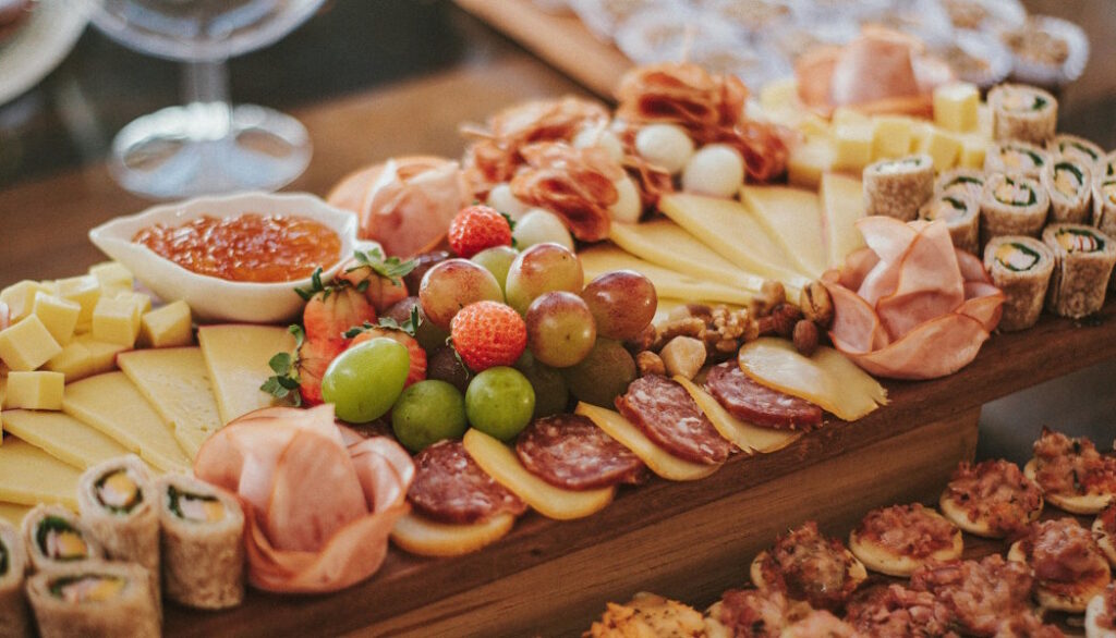 a close-up view of a charuterie board that contains many options such as grapes, cheese, salami circles, strawberries, cured ham roses, pieces of sushi, crackers with cheese and bacon, and many more choices