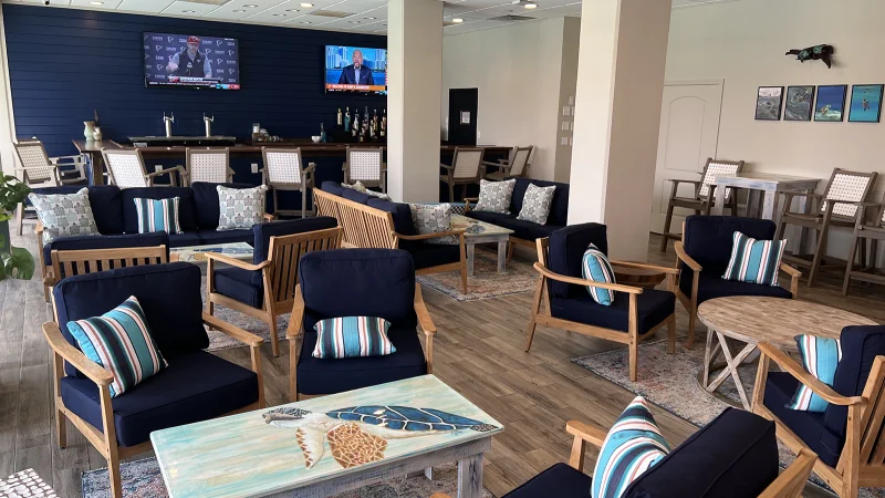 an open spacious room with clustered circles of wooden chairs with navy blue cushions brightly lit by a glass windowed wall with an ocean view, in the background there is a bar with two large TVs over it