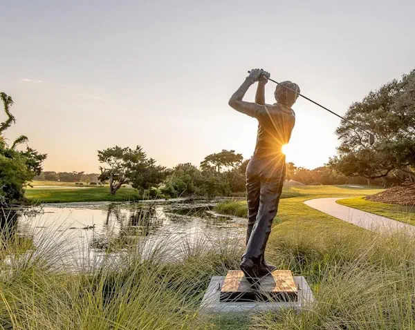 a stone statue of a golfer swinging a golf club, behind it are fields of green grass, a river of water, and a stone pathway leading into the distance.